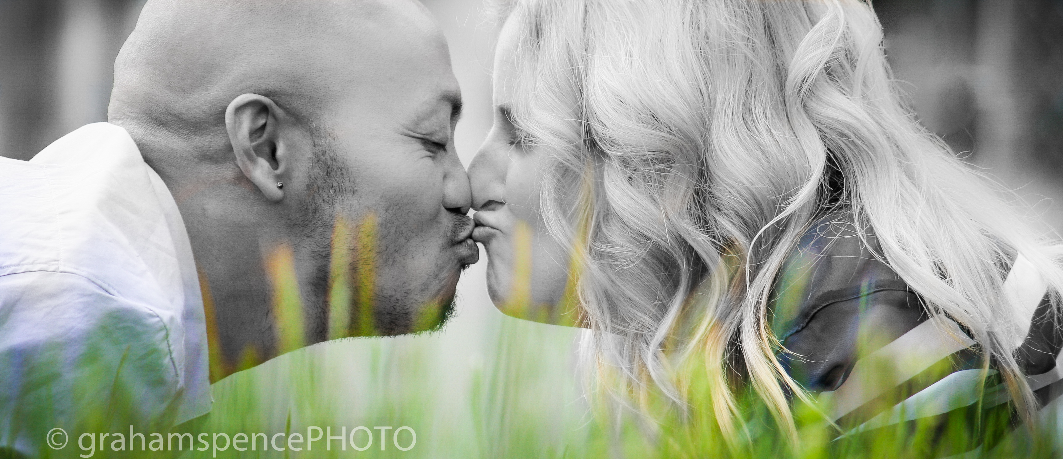 Close-up side view of a recently engaged couple facing each other with a kiss while lying in the grass.